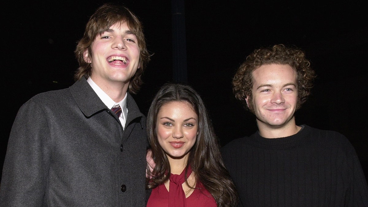 Ashton Kutcher lächelt in Pullover und Krawatte neben Mila Kunis in einem roten Hemd neben Danny Masterson im Jahr 2000