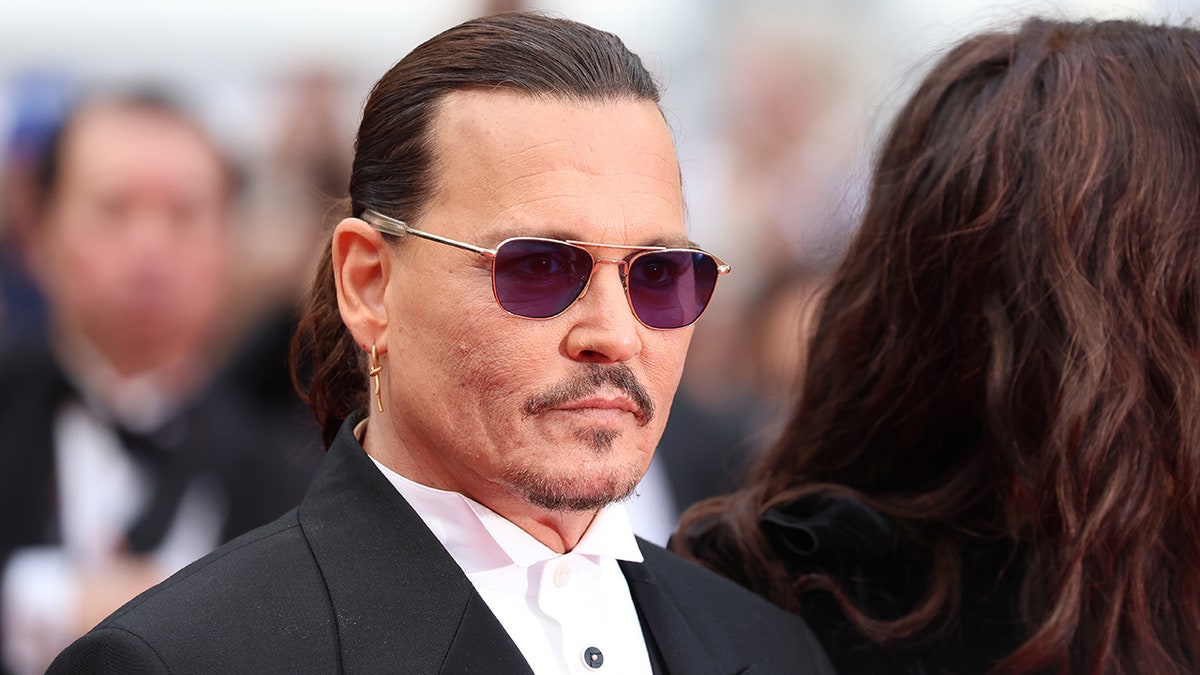 Johnny Depp trägt eine Sonnenbrille auf dem Teppich von Cannes, die Haare nach hinten gekämmt und einen schwarzen Anzug