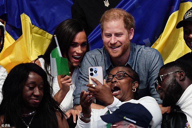 Harry und Meghan lächeln in die Kamera, während ein Fan ein Selfie macht