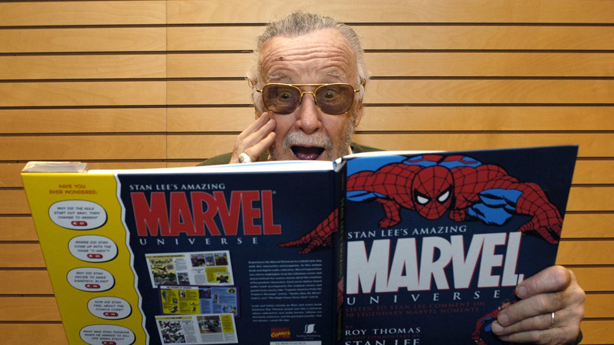 Stan Lee hält sich schockiert die Hand vors Gesicht, während er ein riesiges Marvel-Comic-Buch liest