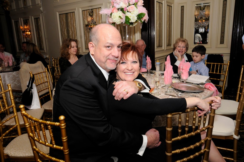 Caroline and Albert Manzo III smiling and embracing while sitting at a table with large flower arrangement and pink napkins. 