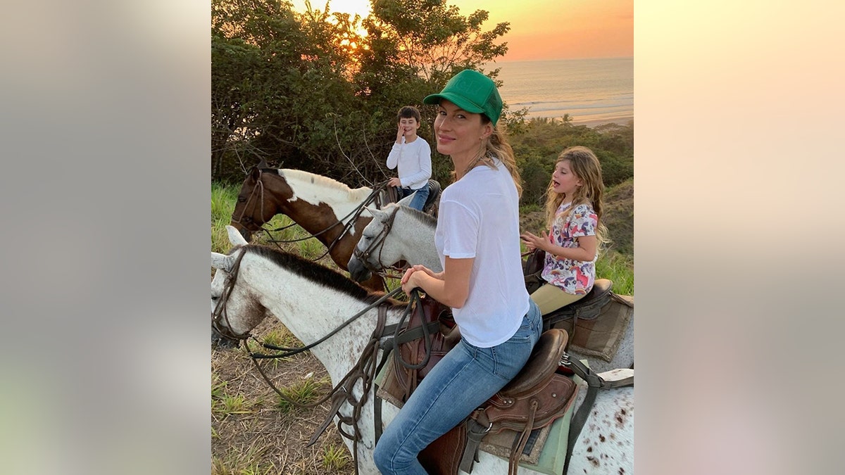 Gisele Bündchen blickt zurück auf ein Foto beim Reiten mit ihren Kindern Benny und Vivi im Hintergrund, ebenfalls auf Pferden und einem Sonnenuntergang im Hintergrund