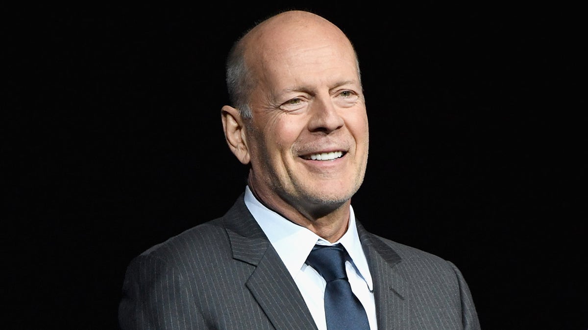 Bruce Willis lächelt auf der Bühne in einem grauen Anzug und einer dunkelblauen Krawatte
