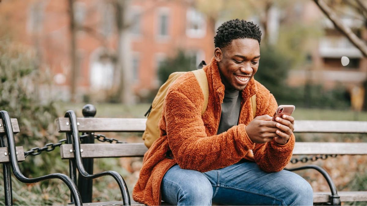 Mann sitzt auf einer Parkbank und lächelt auf sein Telefon.