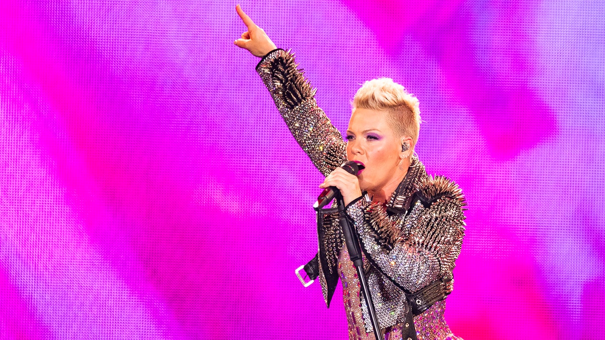 Pink zeigt in einer Lederjacke mit Nieten vor einem magentafarbenen Bildschirm in die Luft und tritt auf der Bühne auf