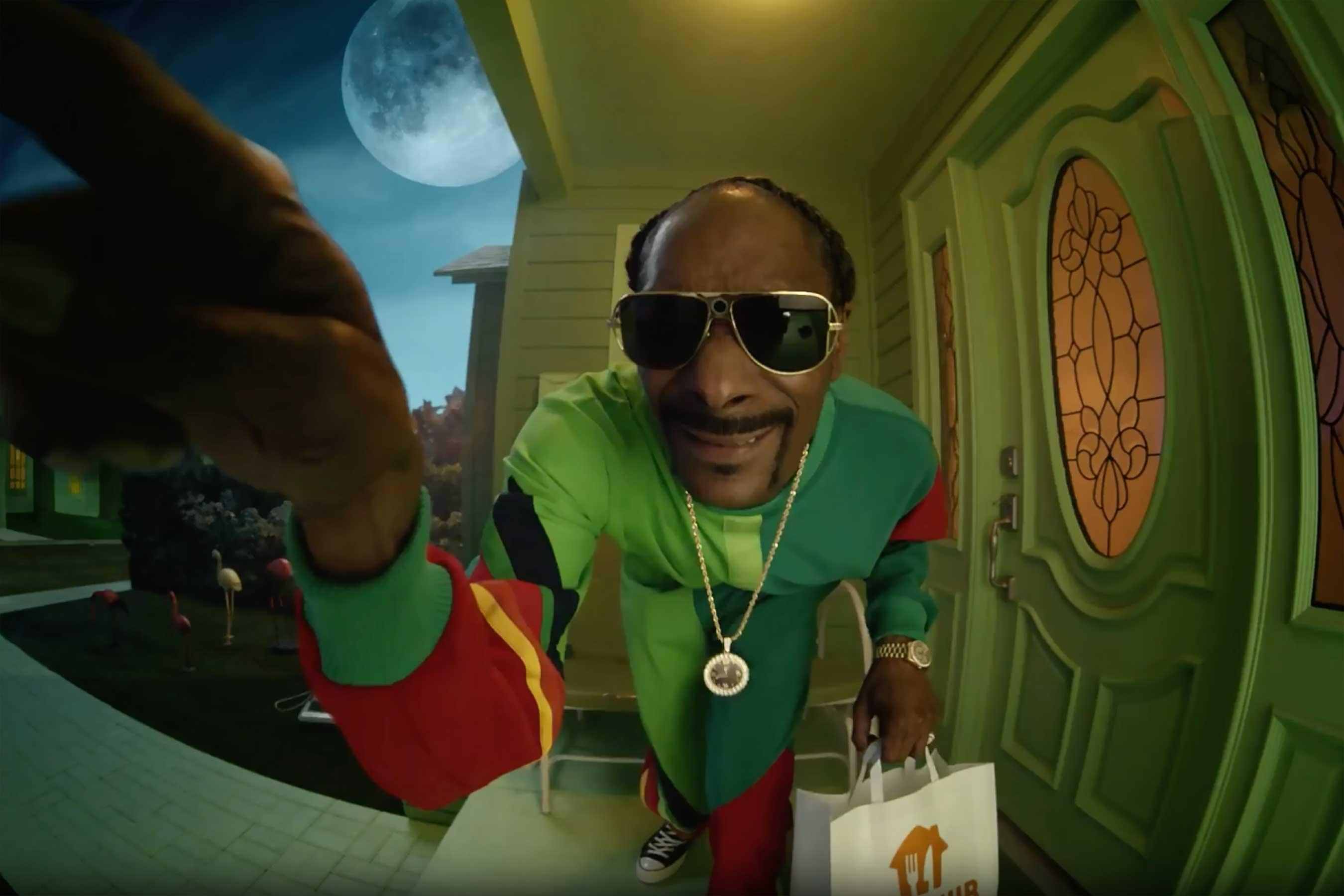 Snoop Dogg trägt einen grün-roten Trainingsanzug und klingelt mit einem weiß-orangefarbenen Hemd an der Türklingel.