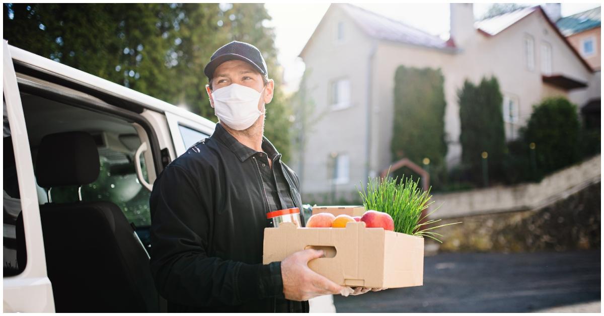Ein männlicher Lieferfahrer mit Lebensmitteln in der Hand und einer Maske.