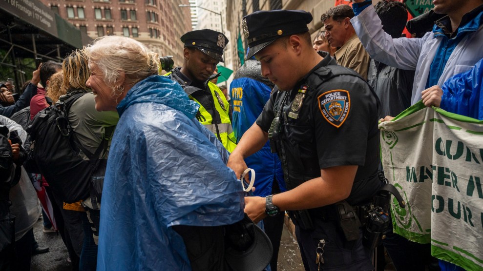 Eine Frau mit weißen Haaren und einem blauen Regenmantel wird von einem NYPD-Beamten festgenommen, im Hintergrund weitere Demonstranten und Polizisten.