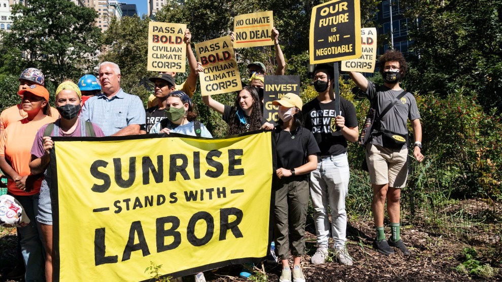 Eine Gruppe von Menschen, größtenteils maskiert, hält gelbe und schwarze Schilder mit Slogans wie „ "Unsere Zukunft ist nicht verhandelbar" Und "Übergeben Sie ein mutiges ziviles Klimakorps." Vor ihnen halten mehrere ein Transparent mit der Aufschrift „ "Sunrise steht mit der Arbeit."