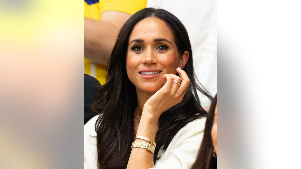 Eine Nahaufnahme von lächelnder Meghan Markle in einem weißen Pullover und einer goldenen Uhr mit passendem Armband