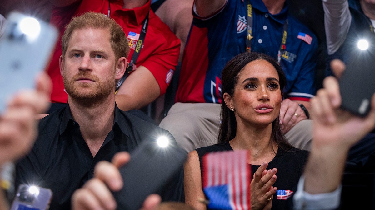 Meghan Markle und Prinz Harry sitzen in einer Menschenmenge, umgeben von Mobiltelefonen