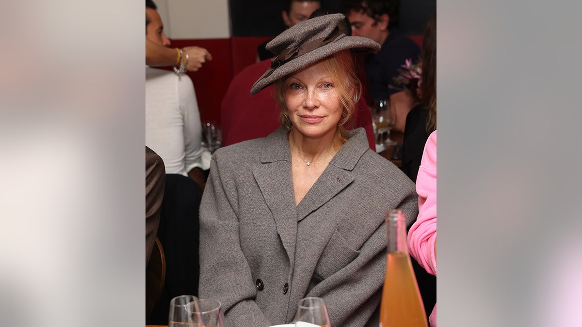 Pamela Anderson sitzt sittsam mit schräg gestelltem Hut auf einer Veranstaltung