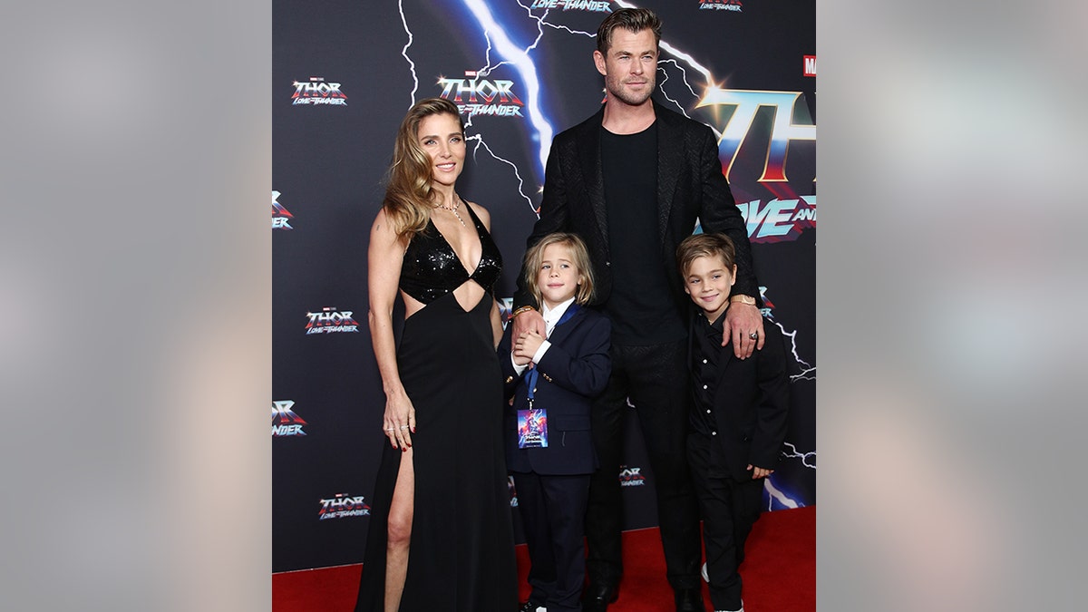 Chris Hemsworth in einem schwarzen Hemd und einer Jacke lächelt mit seinen beiden Söhnen (einer in einem komplett schwarzen Anzug, der andere in einem schwarzen Anzug und einem weißen Hemd) zusammen mit seiner Frau Elsa Pataky in einem schwarzen Kleid mit Ausschnitten auf dem roten Teppich