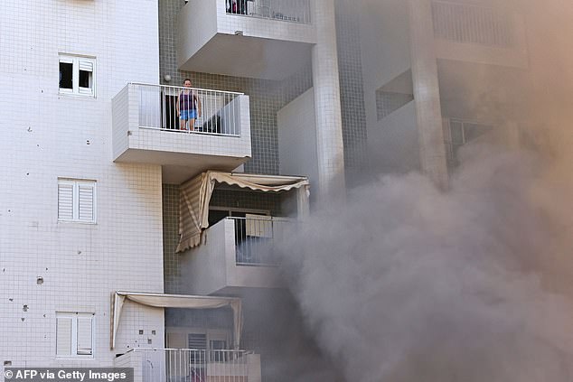 Eine Frau steht auf ihrem Balkon, während nach einem Raketenangriff aus dem Gazastreifen in der südisraelischen Stadt Aschkelon Rauch auf dem Parkplatz eines Wohngebäudes aufsteigt