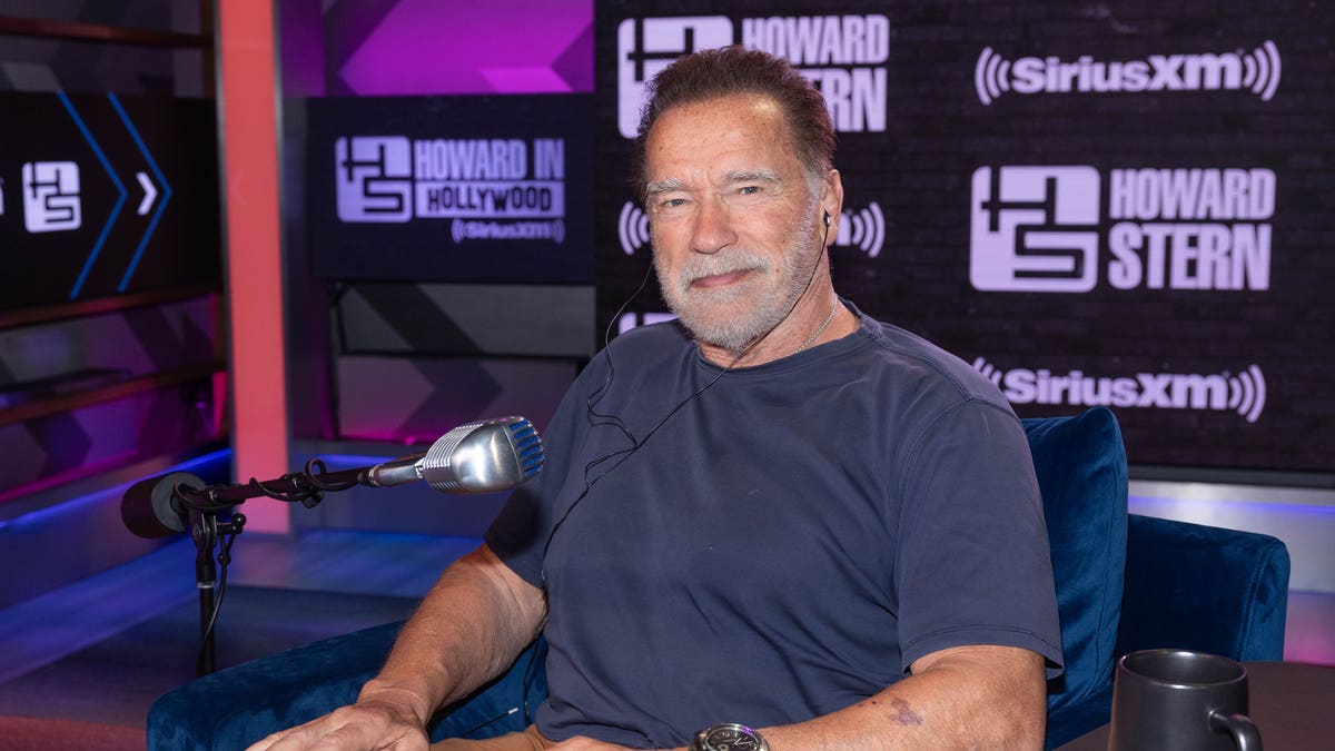 Arnold Schwarzenegger im blauen Hemd setzt sich an einen Tisch "Die Howard Stern Show"