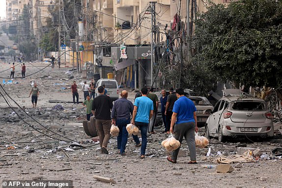 Palestinian men carry bread through a heavily bombed street following Israeli airstrikes on Gaza City on October 10, 2023. Israel kept up its deadly bombardment of Hamas-controlled Gaza on October 10 after the Palestinian militant group threatened to execute some of the around 150 hostages it abducted in a weekend assault if air strikes continue without warning. (Photo by MAHMUD HAMS / AFP) (Photo by MAHMUD HAMS/AFP via Getty Images)
