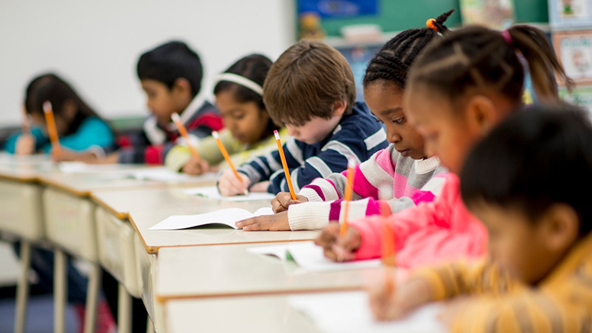 Eine multiethnische Gruppe von Grundschulkindern sitzt drinnen in einem Klassenzimmer.  Sie tragen Freizeitkleidung.  Die Schüler sitzen an ihren Schreibtischen und schreiben mit Bleistiften.