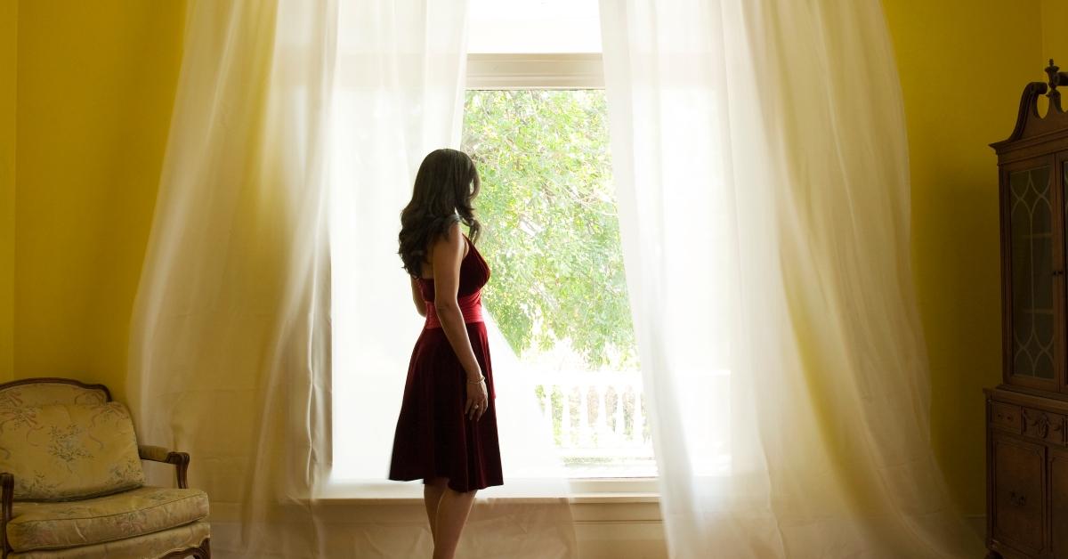 Eine Frau steht draußen und blickt auf ein großes offenes Fenster mit wehenden Vorhängen