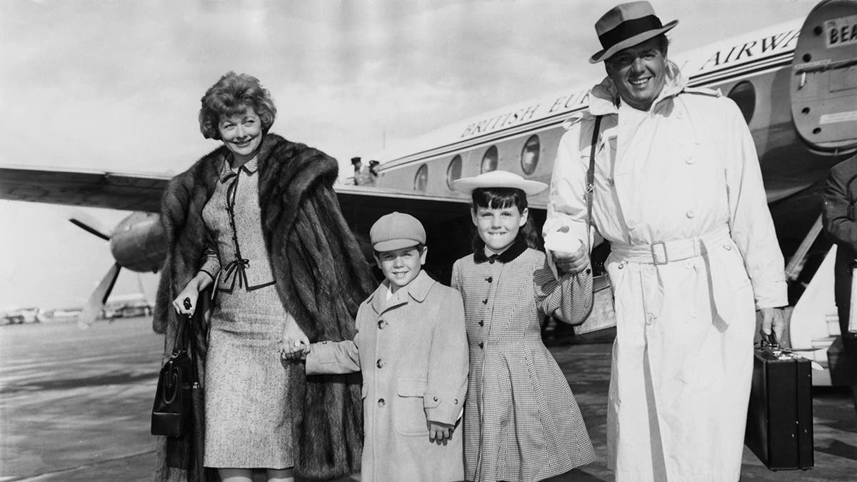 Lucille Ball und Desi Arnaz tragen Mäntel, während sie ihre beiden kleinen Kinder vor einem Flugzeug festhalten