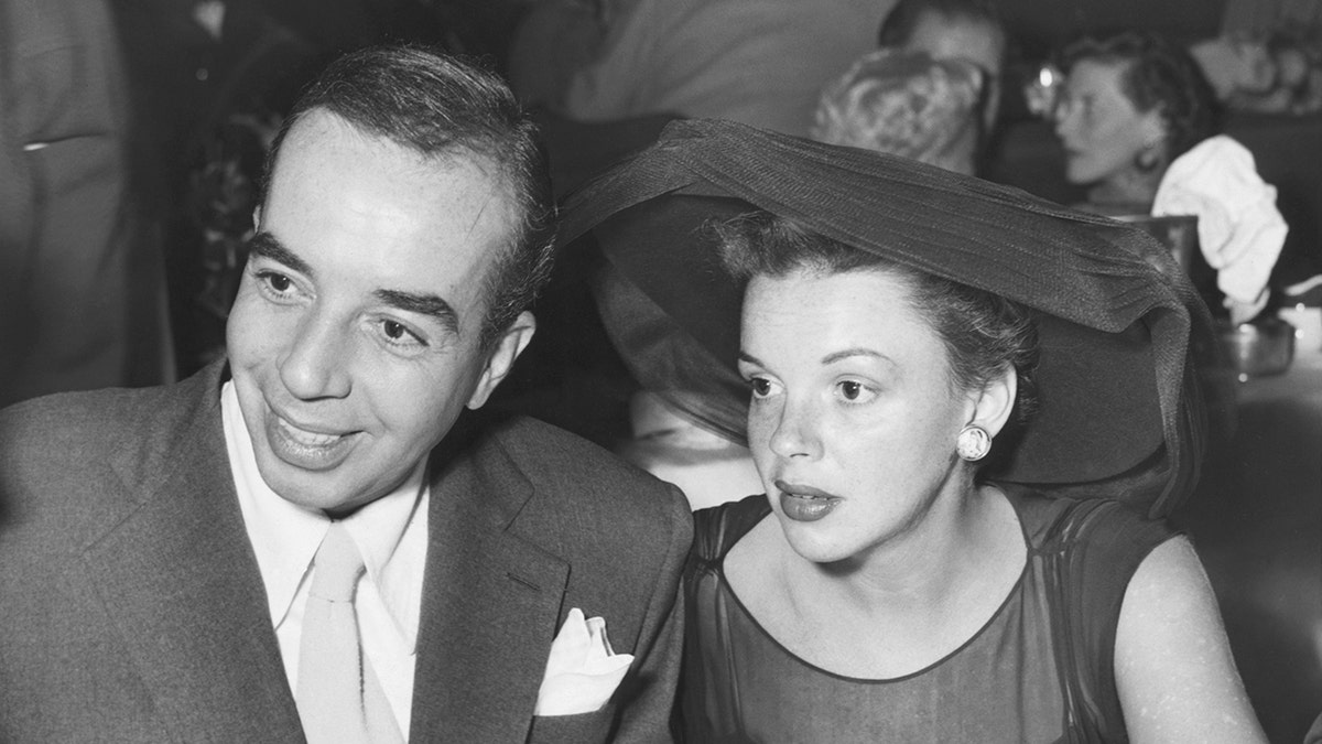 Judy Garland sieht ernst aus in einem schwarzen Kleid und passendem Hut neben ihrem Mann