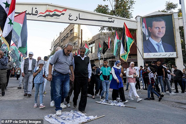 People walk on makeshift Israeli flags strewn on the ground past Palestinian members of the Saraya al-Quds, the military branch of Islamic Jihad faction, standing in line holding Palestinian flags during a rally organised by their faction at the Yarmouk camp for Palestinian refugees south of Syria's capital Damascus