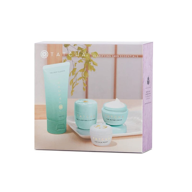 Tatcha Skin Essentials Set: Eine Schachtel auf weißem Hintergrund mit dem Bild einer hellblauen Tube, zwei kleine passende Gläser und ein kleines weißes Glas, das auf einem Holztisch mit einem Fenster vor einem Fenster steht