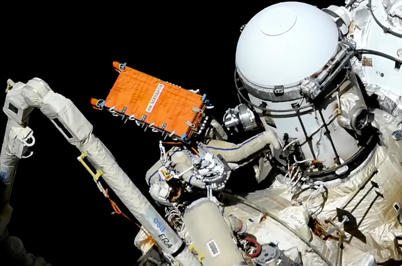 Ein Astronaut im weißen Raumanzug installiert ein orangefarbenes Radarkommunikationssystem auf der internationalen Raumstation.