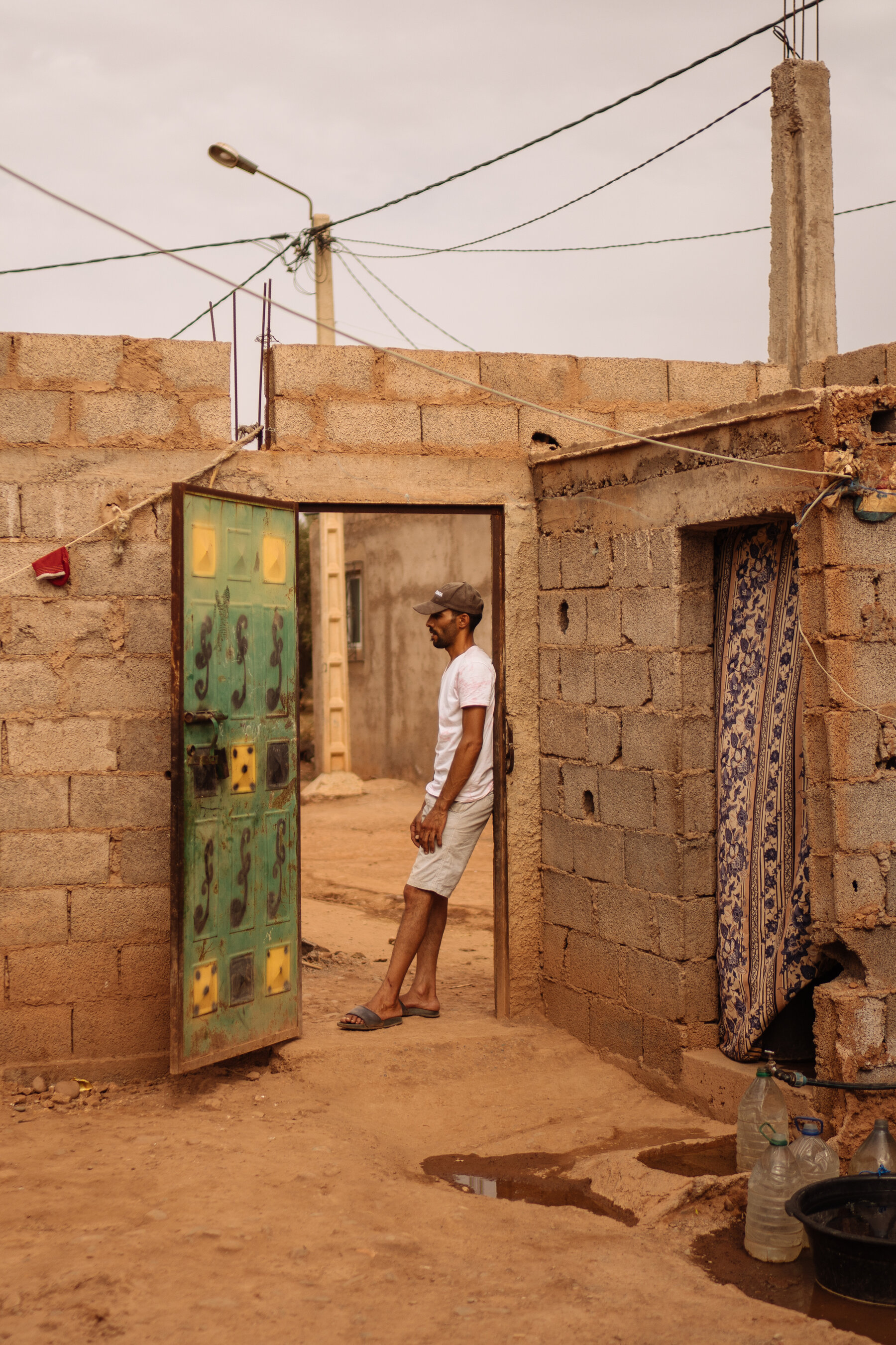 A man in a white t-shirt and shorts leaning against the open doorway of a cinderblock house.