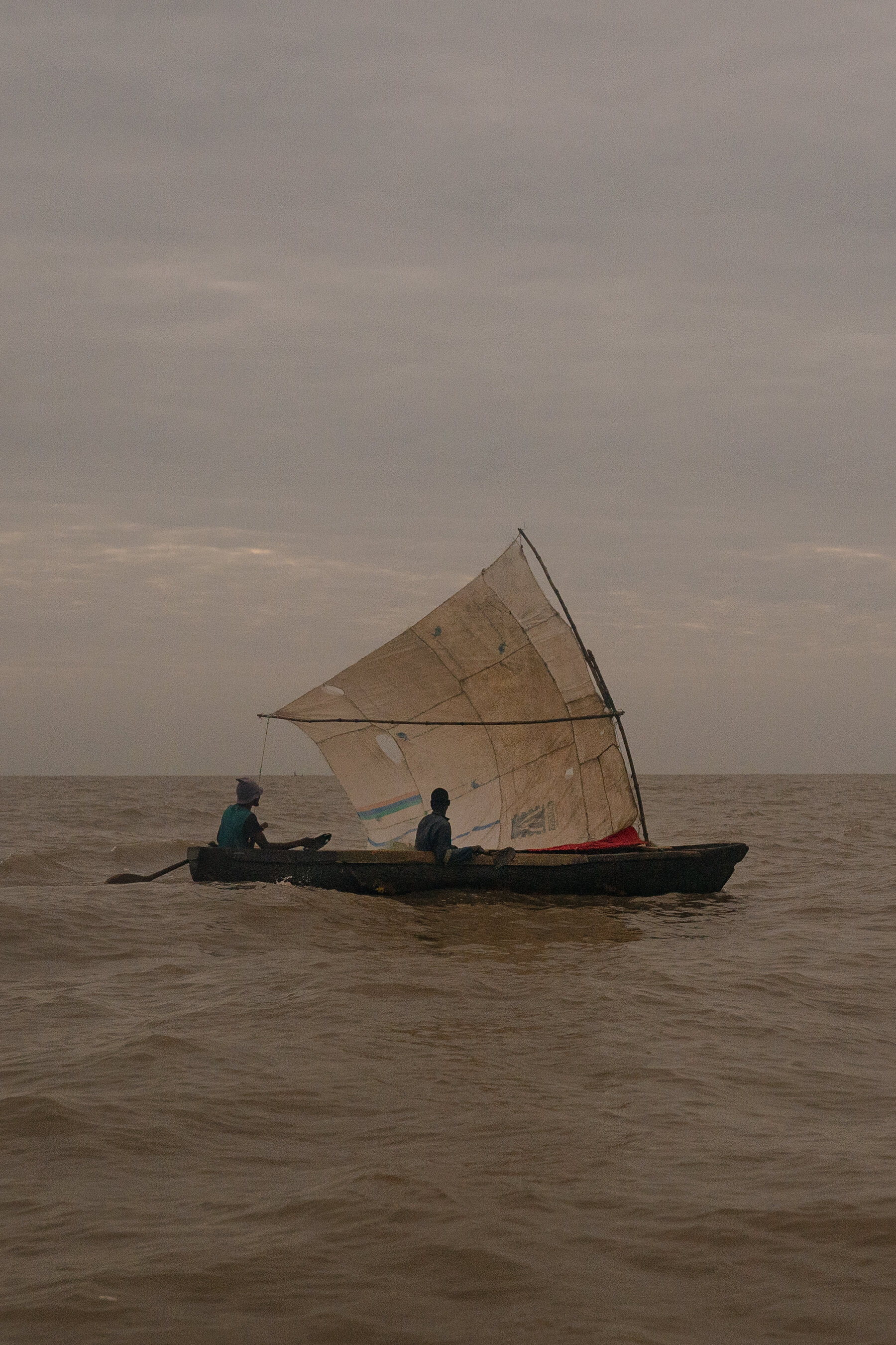 Two people on the open sea in a small boat with a tattered sail.