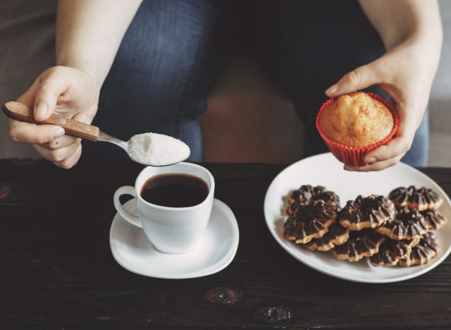 Frau gießt Zucker in Kaffee und hält Muffin über Teller mit Keksen