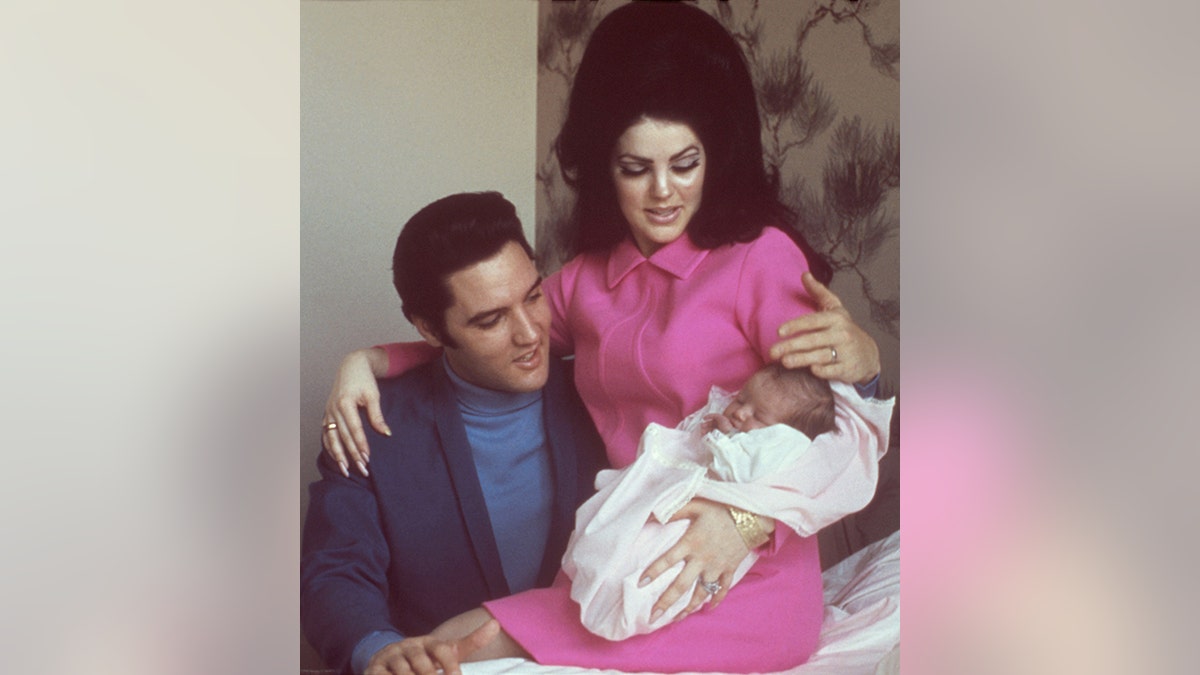 Elvis Presley in blauem Hemd und dunkelblauer Jacke betrachtet Lisa Marie Presley als Baby, das von Priscilla Presley in Rosa gehalten wird