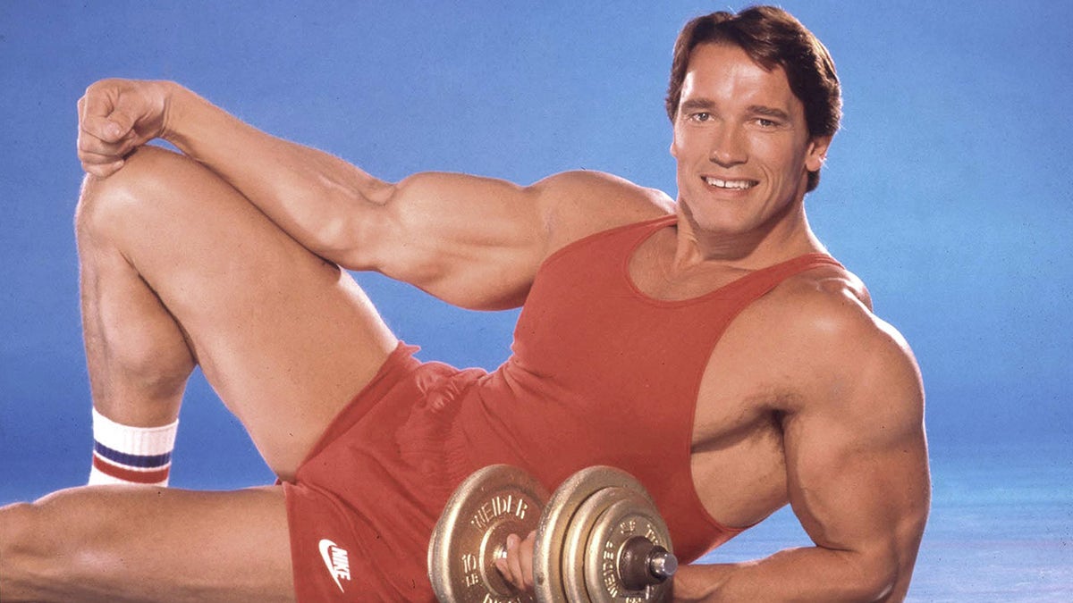 Arnold Schwarzenegger hält Hanteln, während er in roter Fitnessausrüstung posiert