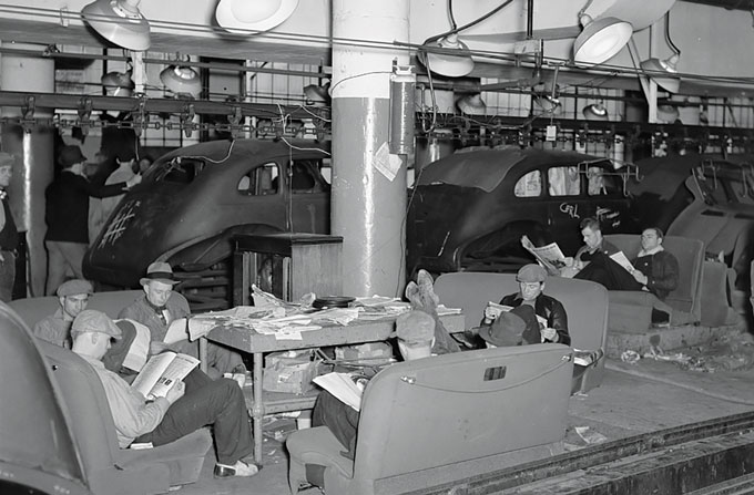 Workers sit reading papers on couches in a factory setting with partially built automobiles in the background.