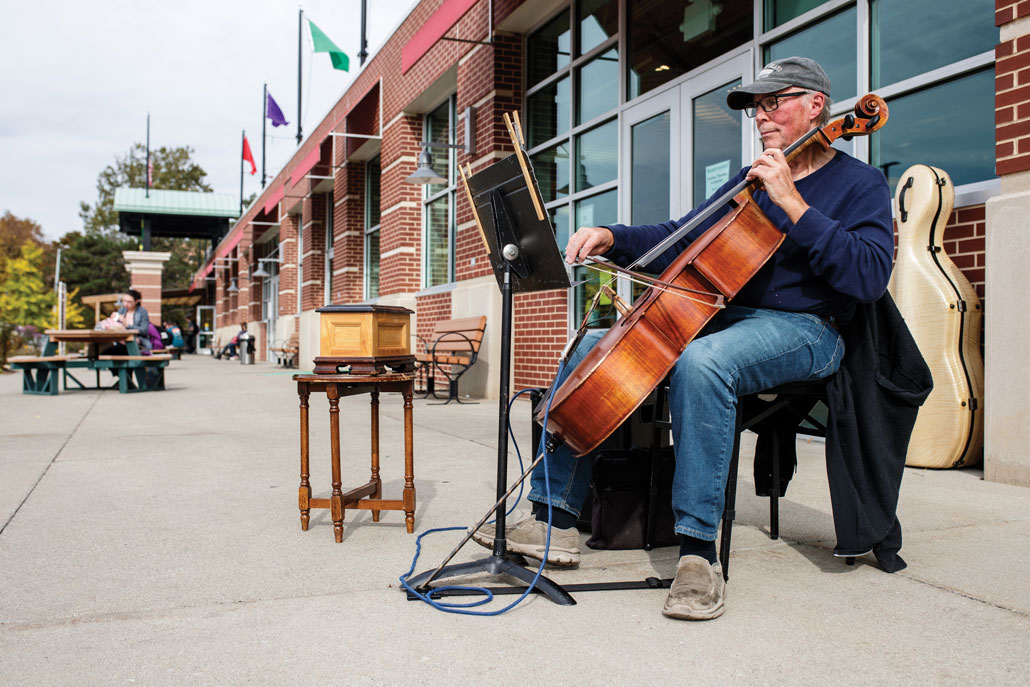 A person plays the cello on a chair outside a brick building.