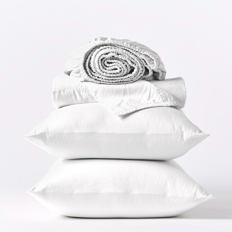 Coyuchi Cloud Weiches Bio-Satin-Bettlaken-Set: Ein weißes gefaltetes Spannbettlaken, ein passendes gefaltetes Flachlaken und zwei übereinander gestapelte Kissen auf grauem Hintergrund