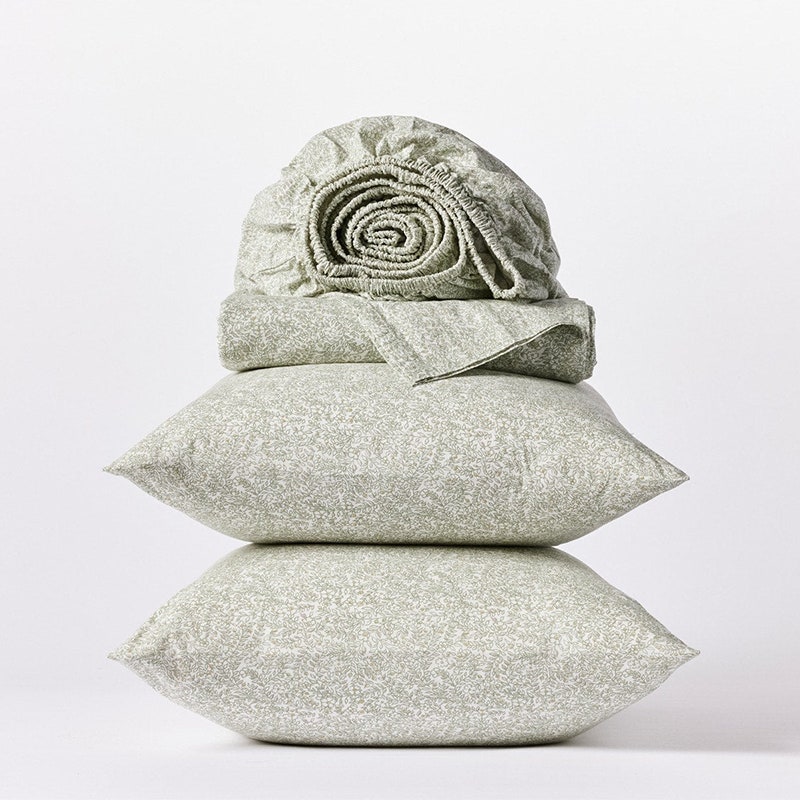 Coyuchi Organic Print Perkal-Bettlaken-Set: Ein gefaltetes Spannbettlaken in Heidegrau, ein passendes gefaltetes Flachlaken und zwei übereinander gestapelte Kissen auf grauem Hintergrund