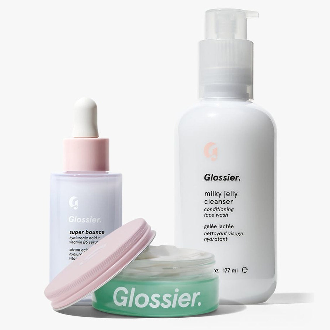 Glossier The Winter Skin Set: grünes Glas, lila Serumflasche, weiße Pumpflasche auf grauem Hintergrund