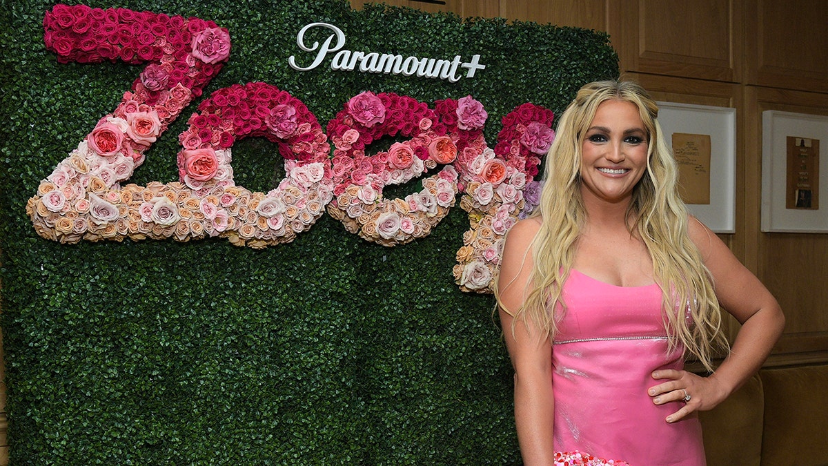 Jamie Lynn Spears in einem rosa Kleid legt ihre Hand in die Hüfte und steht vor einer grünen Wand, auf der Zoey in Blumen geschrieben steht
