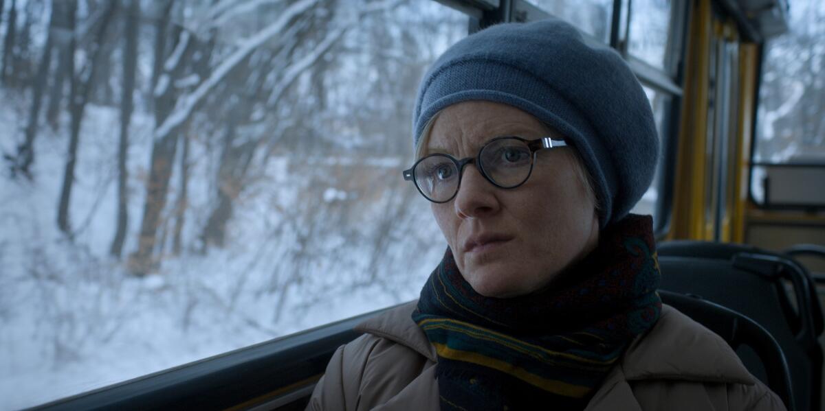 Eine Frau mit grauer Mütze, Schal und Brille sitzt in einem Auto, das an einem verschneiten Wald vorbeifährt.