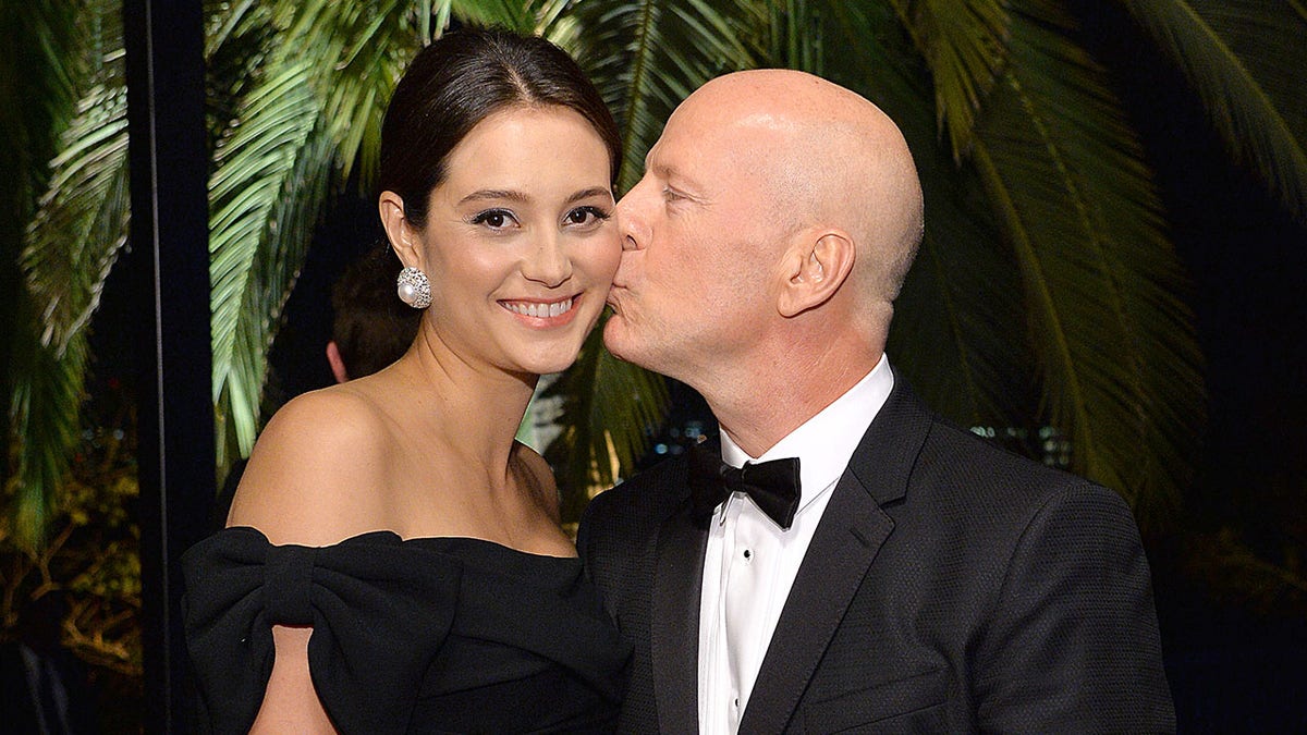 Bruce Willis im klassischen Smoking küsst seine Frau Emma Heming Willis in einem schwarzen Kleid auf die Wange