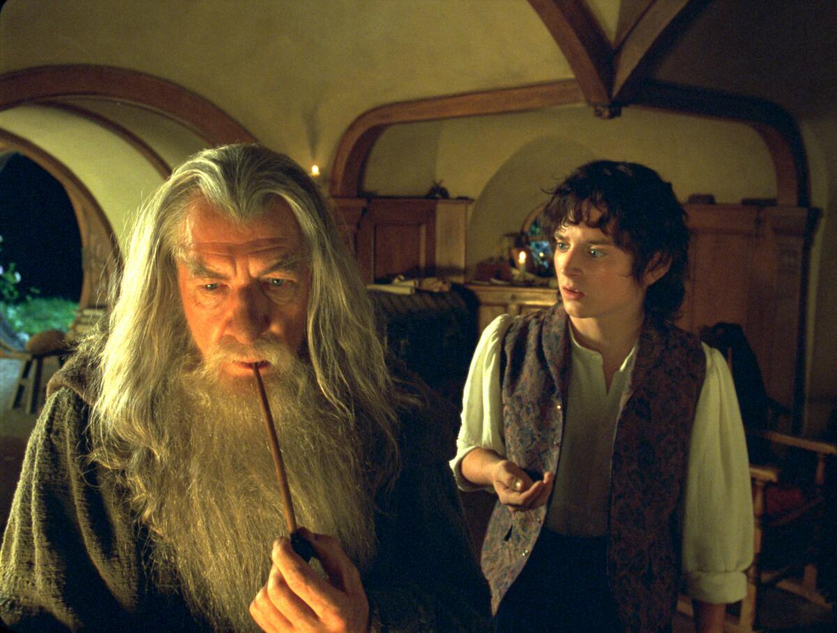 Ein junger Hobbit steht hinter einem Zauberer mit langen grauen Haaren und Bart, der eine Pfeife raucht