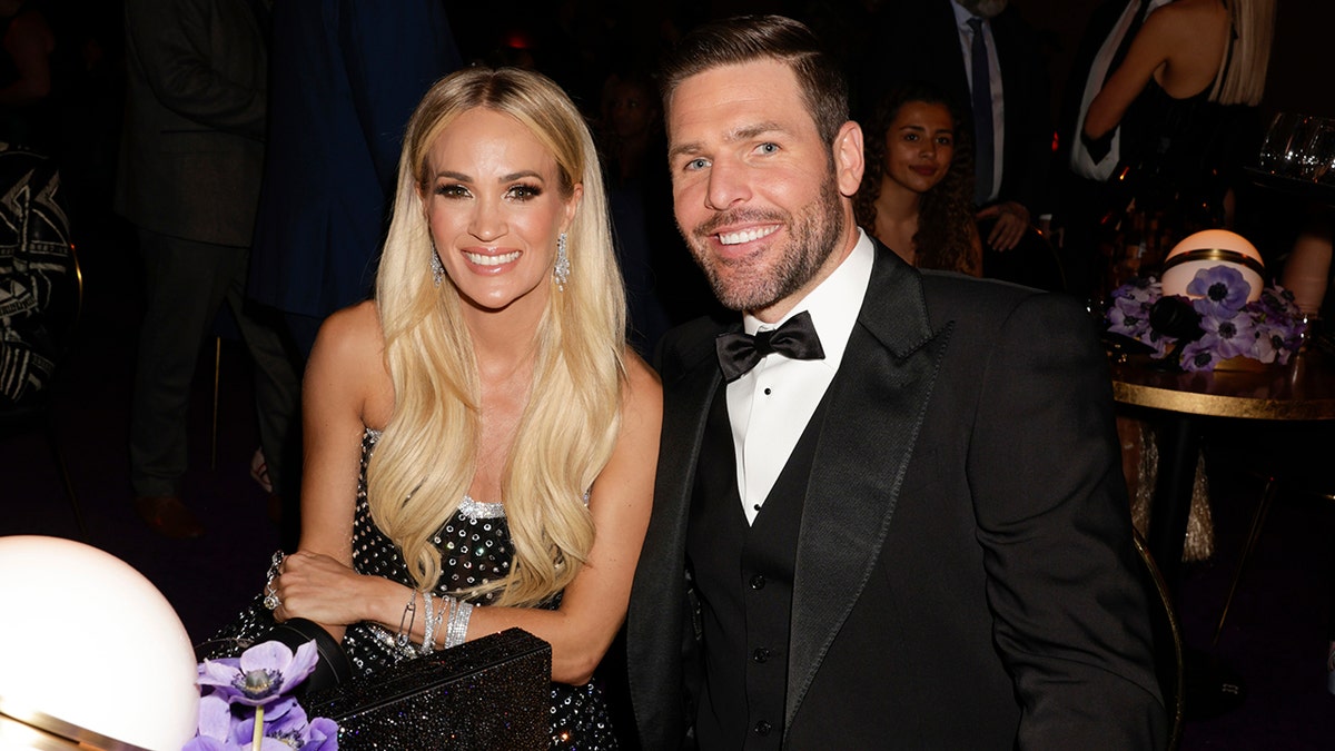 Carrie Underwood mit langen blonden Haaren sitzt neben ihrem Mann Mike Fisher im Smoking an einem Tisch bei der Grammy-Verleihung
