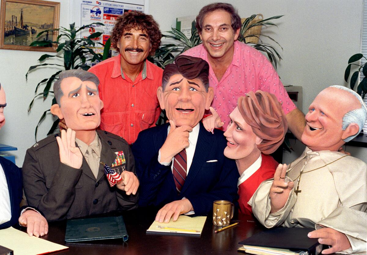 Sid Krofft und Marty Krofft stehen hinter vier lebensgroßen Puppen in Anzügen, die an einem Tisch sitzen.