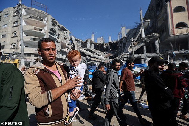 A man holds a child as Palestinians shop in an open-air market near the ruins of houses and buildings destroyed in Israeli strikes in Nuseirat refugee camp in the central Gaza Strip on Thursday