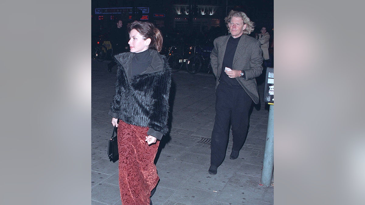 Shania Twain in einem rot gemusterten Samtrock, einem schwarzen Top und einer schwarzen Jacke geht vor Ehemann Robert John Mutt Lange in schwarzen Hosen und einer grauen Jacke her