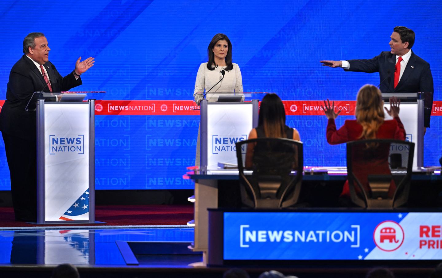 Auf der Debattenbühne gestikulieren und zeigen Chris Christie und Ron DeSantis aufeinander, während Nikki Haley zwischen ihnen zuschaut.