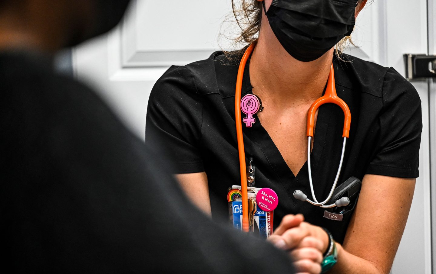 Eine Ärztin in einem schwarzen Kittel und einer schwarzen Maske, trägt eine Regenbogennadel und eine Frauennadel, mit einem Stethoskop um den Hals, und umklammert die Hände eines Patienten.