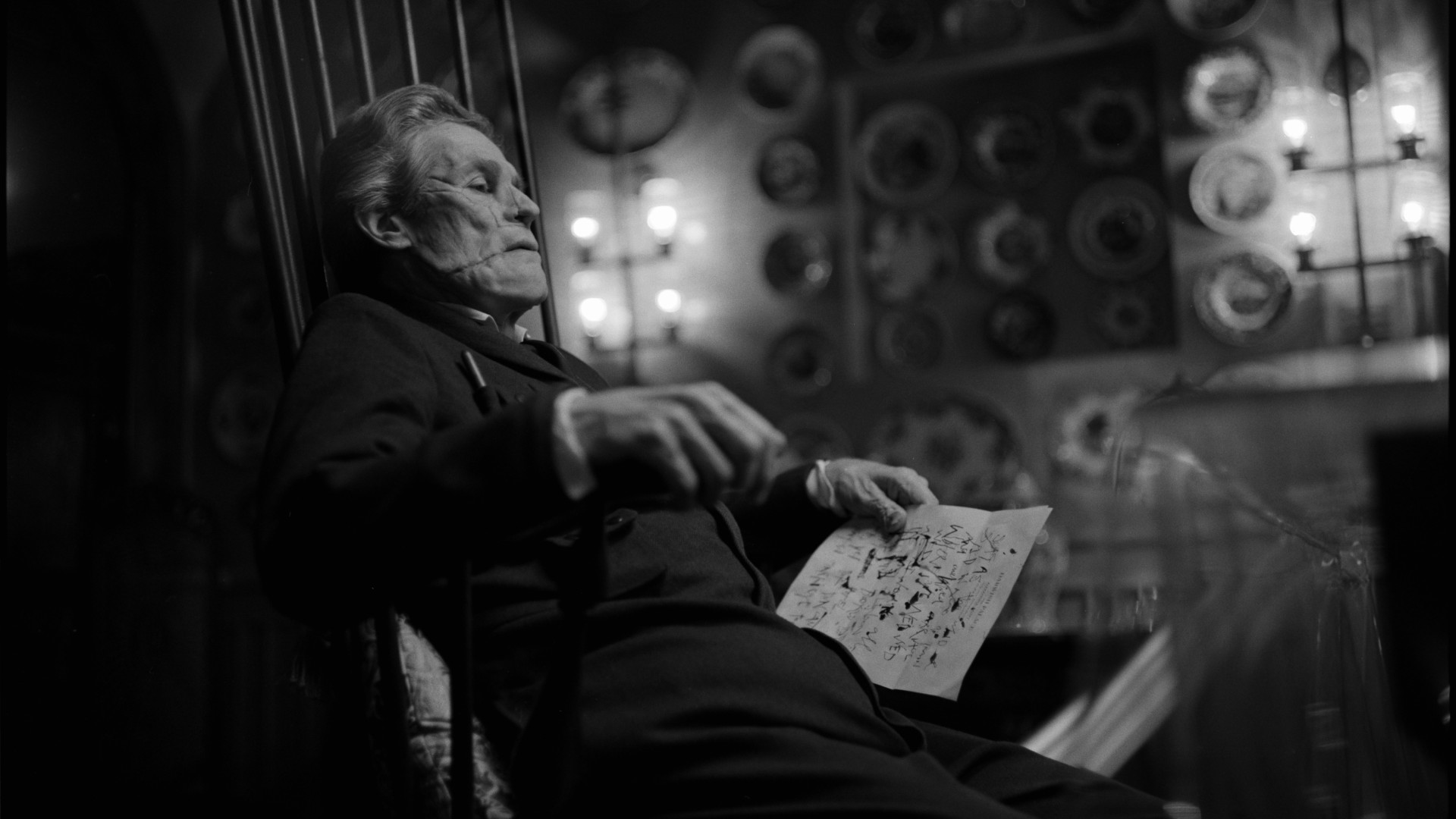 Ein deformierter Willem Dafoe sitzt auf einem Stuhl und hält Papiere in einem Schwarz-Weiß-Standbild von Poor Things in der Hand.