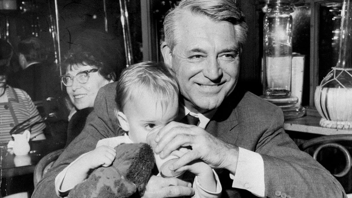 Cary Grant lächelt und hält seine Tochter Jennifer fest, während sie aus einer Tasse trinkt