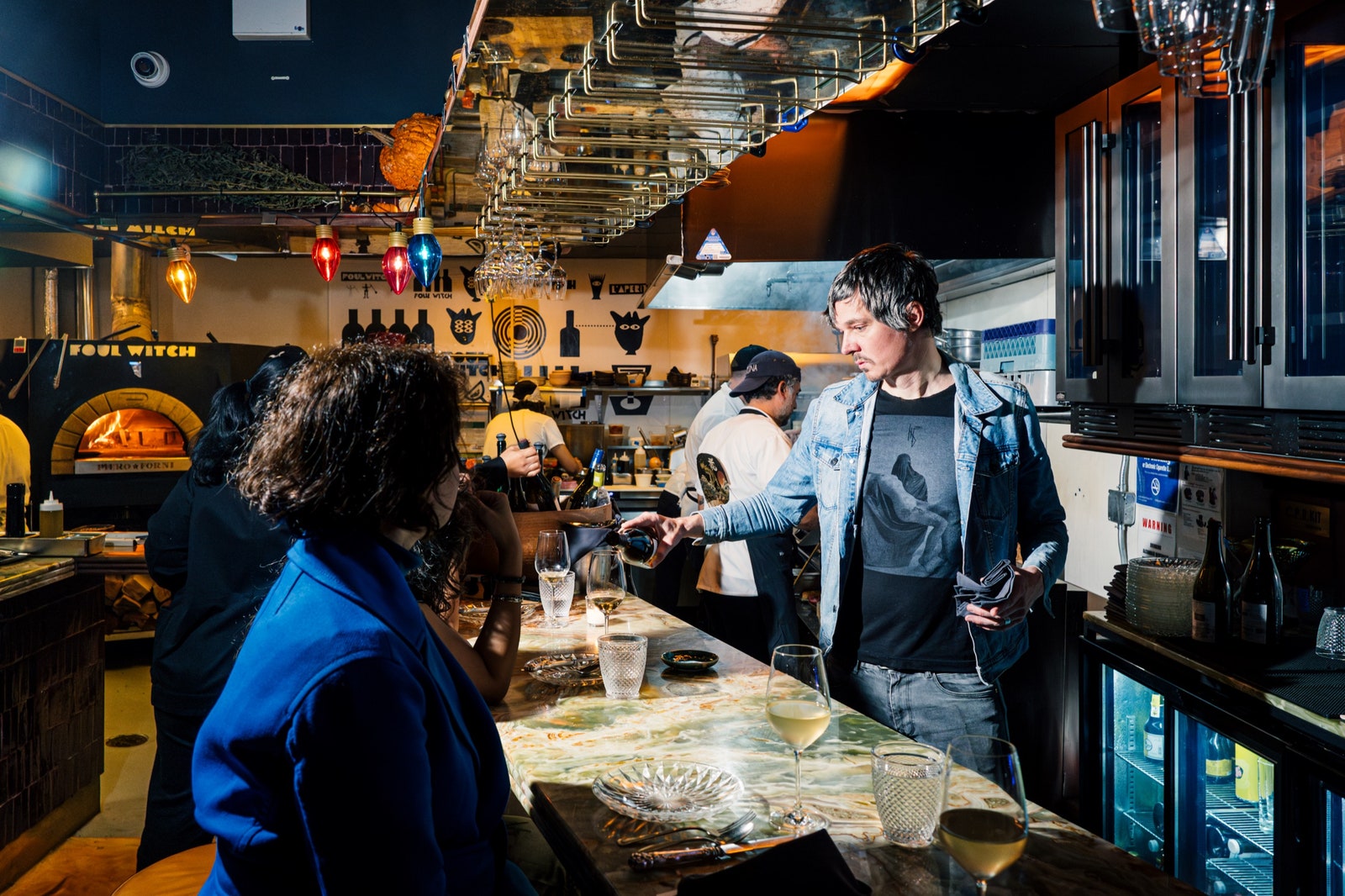 Eine Person serviert den Gästen Wein an einer Bar, im Hintergrund ist eine offene Küche zu sehen.